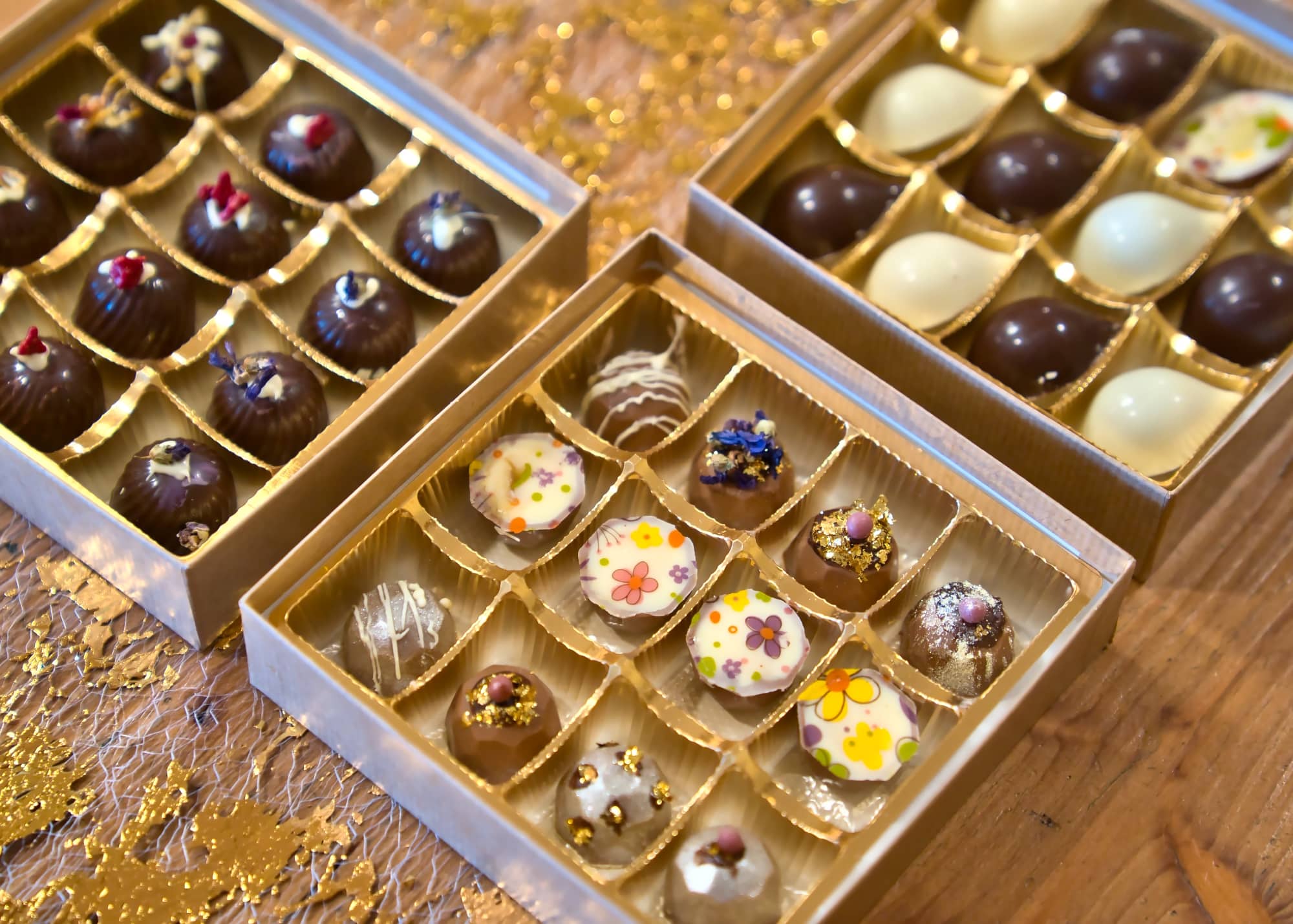 Drei edle Pralinenschachteln mit kunstvoll verzierten Pralinen auf einem festlich dekorierten Holztisch. Die Pralinen sind liebevoll mit Blüten, Goldflocken, Mustern und farbigen Überzügen gestaltet. In einer Schachtel dominieren florale Dekore auf weißer Schokolade, in der anderen glänzen dunkle Pralinen mit feinen Toppings wie Trockenblüten und Streuseln. Die dritte Box enthält eine Mischung aus weißen und dunklen Halbkugelpralinen. Goldene Dekoelemente und ein strukturiertes Netzgewebe unterstreichen den handgefertigten, luxuriösen Charakter der Pralinen.