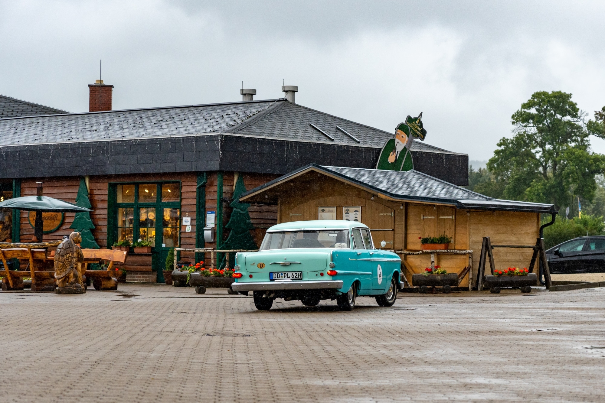 Ein türkis-weißer Oldtimer mit der Startnummer 44 und dem H-Kennzeichen „DD PL 62H“ steht auf einem nassen Platz vor einem rustikalen Gebäude mit Holzfassade und grünem Fensterrahmen. Auf dem Dach ragt eine Figur eines bärtigen Mannes mit grünem Mantel und Zipfelmütze hervor – vermutlich eine Werbefigur für die Einrichtung. Im Vordergrund stehen Holzbänke und geschnitzte Holzfiguren, darunter eine Schildkröte. Der Himmel ist bewölkt, es regnet leicht und die Szene wirkt herbstlich und ländlich.