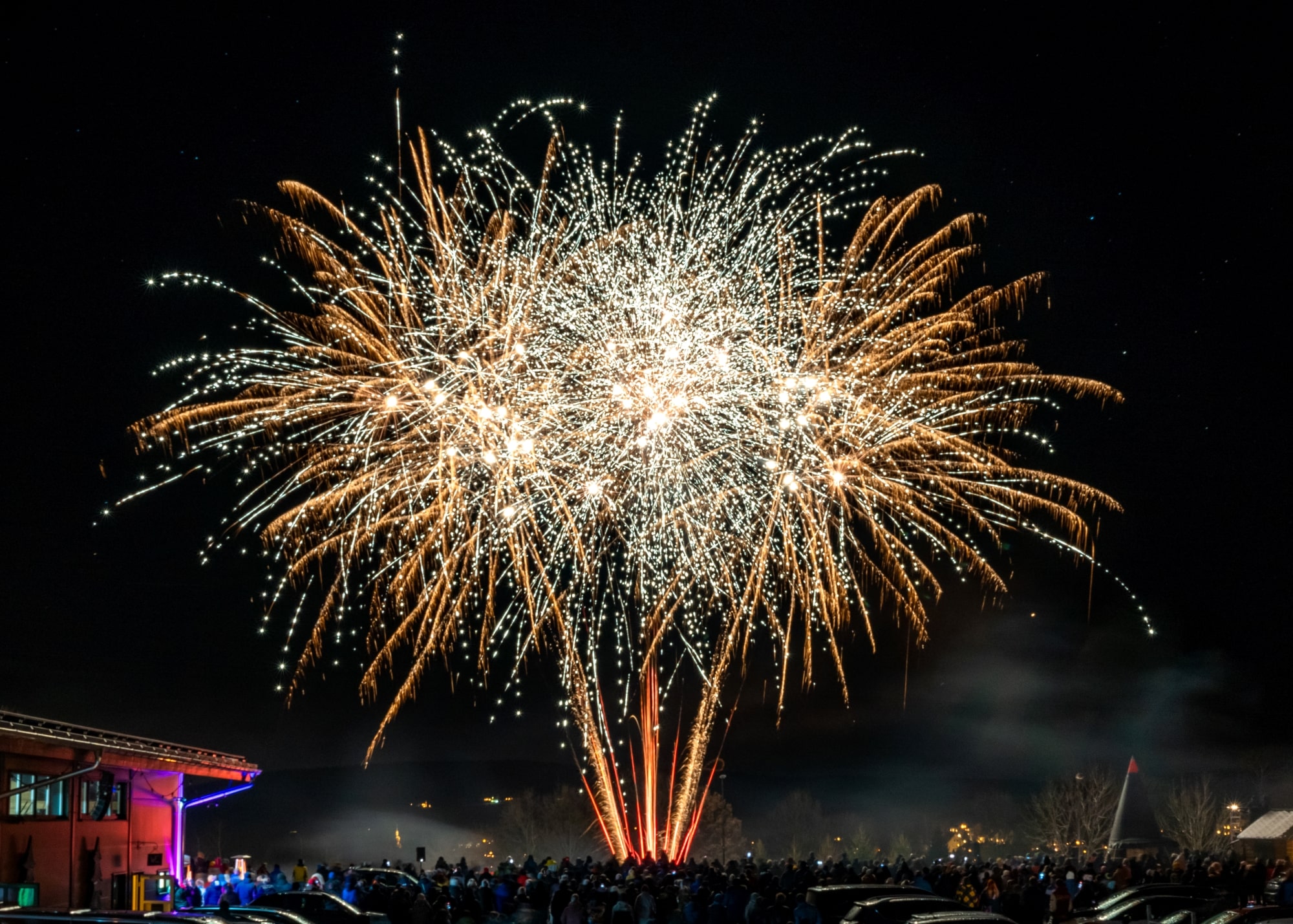 Ein großes, goldenes Feuerwerk explodiert in funkelnden Sternen am Nachthimmel über einer Menschenmenge. Die Zuschauer versammeln sich vor einem Gebäude mit bunter Beleuchtung auf der linken Seite. Rechts im Bild sind geparkte Autos, eine Hütte in Räucherkerzenform und beleuchtete Bäume zu sehen. Der Himmel ist dunkel und klar, einige Sterne sind sichtbar.