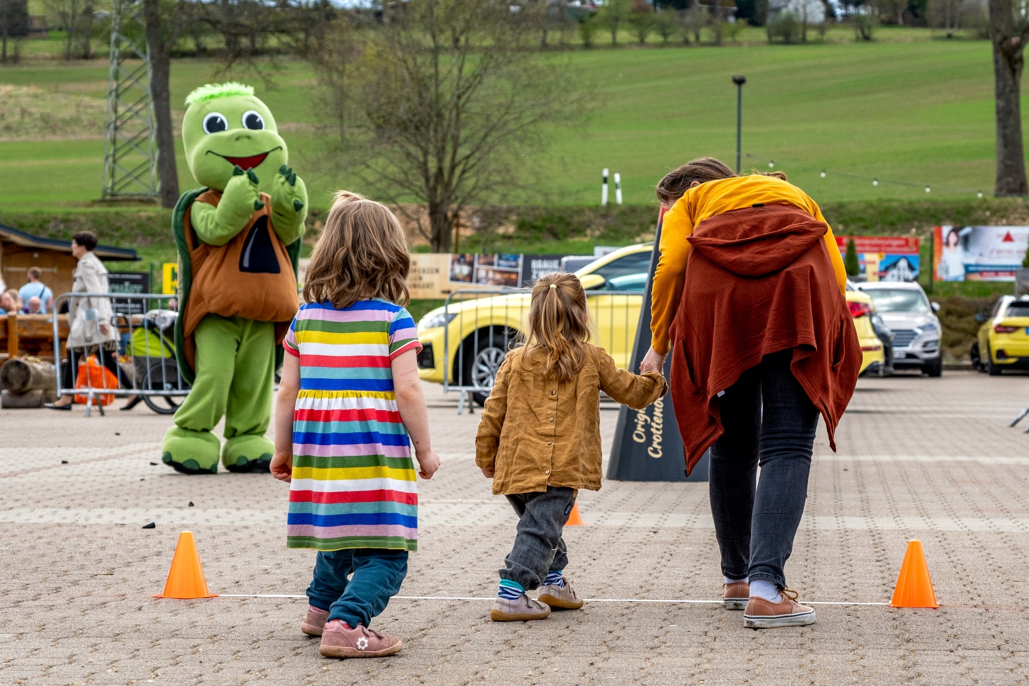 Zwei kleine Kinder und eine erwachsene Person gehen gemeinsam über einen mit Hütchen markierten Parcours auf einem gepflasterten Platz. Im Hintergrund begrüßt ein großes grünes Maskottchen in Schildkrötenkostüm freundlich die Besucher. Um sie herum sind parkende Autos, weitere Menschen und grüne Wiesen im Hintergrund zu sehen.