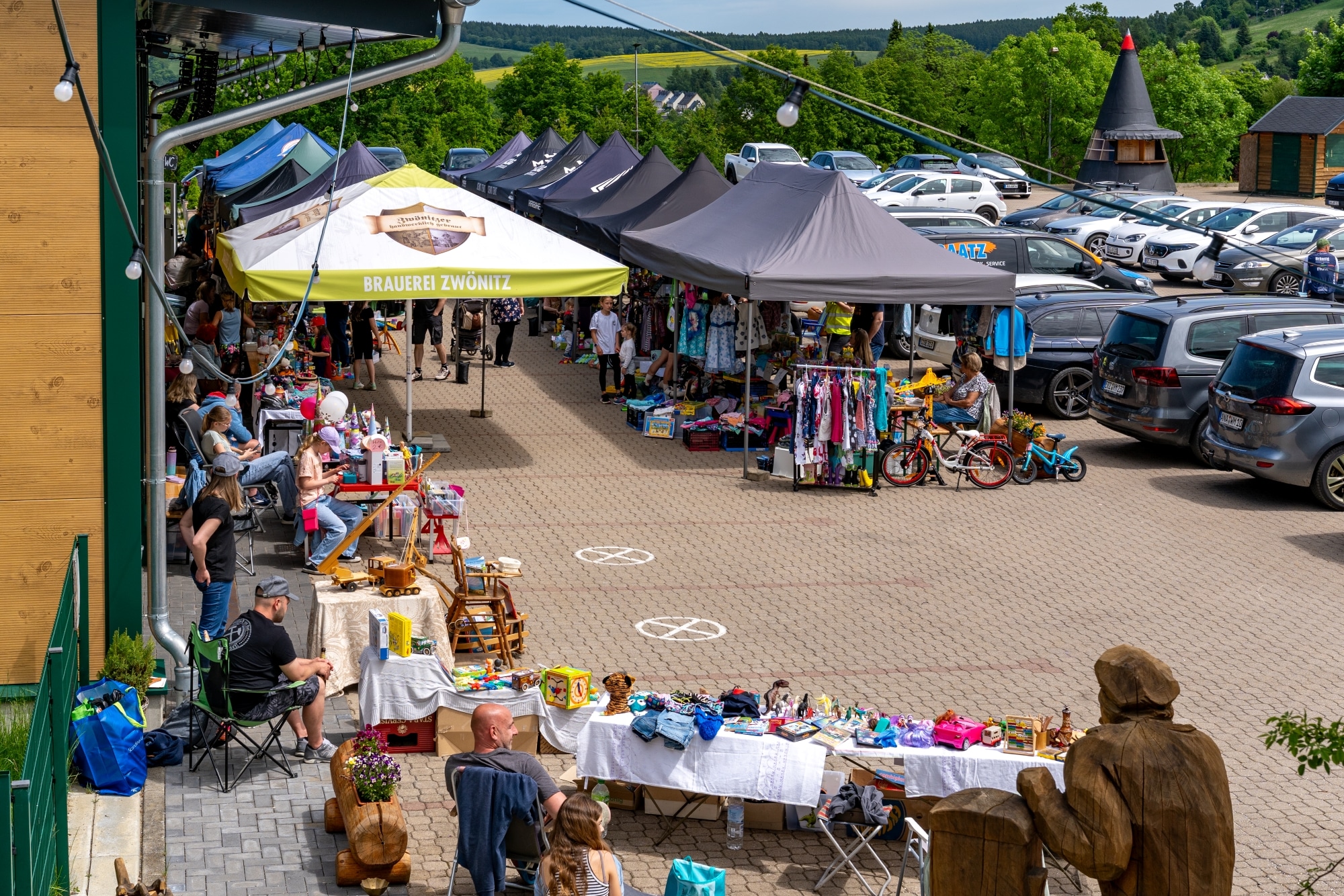 Ein Kinderflohmarkt mit mehreren Ständen, die unter Pavillons stehen. Auf den Tischen werden Kleidung, Spielzeug und Haushaltsgegenstände präsentiert. Menschen schlendern durch die Reihen. Im Hintergrund sind geparkte Autos und eine grüne Landschaft sichtbar.