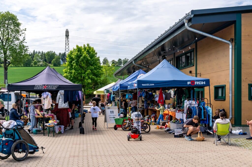 Eine Nahaufnahme eines Flohmarktbereichs mit blauen und schwarzen Pavillons. Kleidung, Spielzeug und Kinderfahrräder werden zum Verkauf angeboten. Im Hintergrund ist eine grüne Wiese zu sehen.