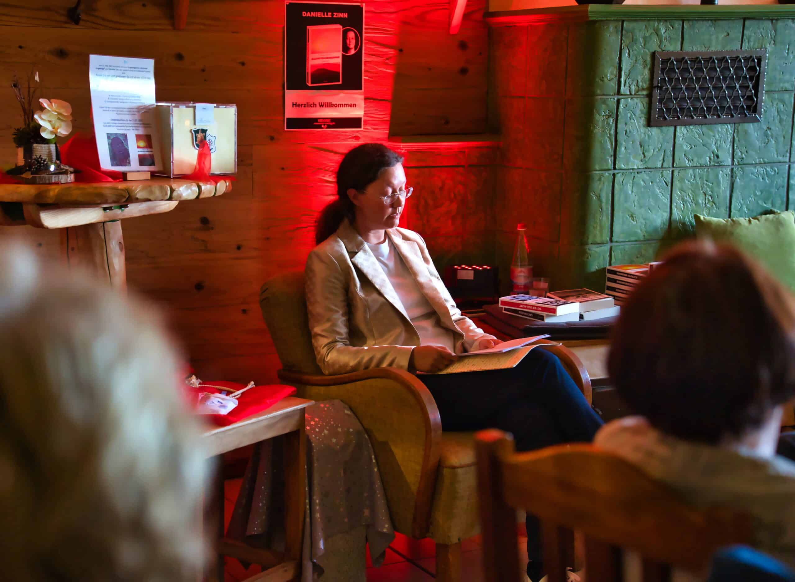 Danielle Zinn in einem hellen Blazer sitzt in einem gemütlichen Holzraum und liest aus einem Buch vor. Im Hintergrund ist ein rotes Poster zu sehen, das auf eine Lesung mit dem Titel „Glutrotes Erzgebirge“ hinweist. Neben ihr befinden sich Bücher und Getränke auf einem Tisch.