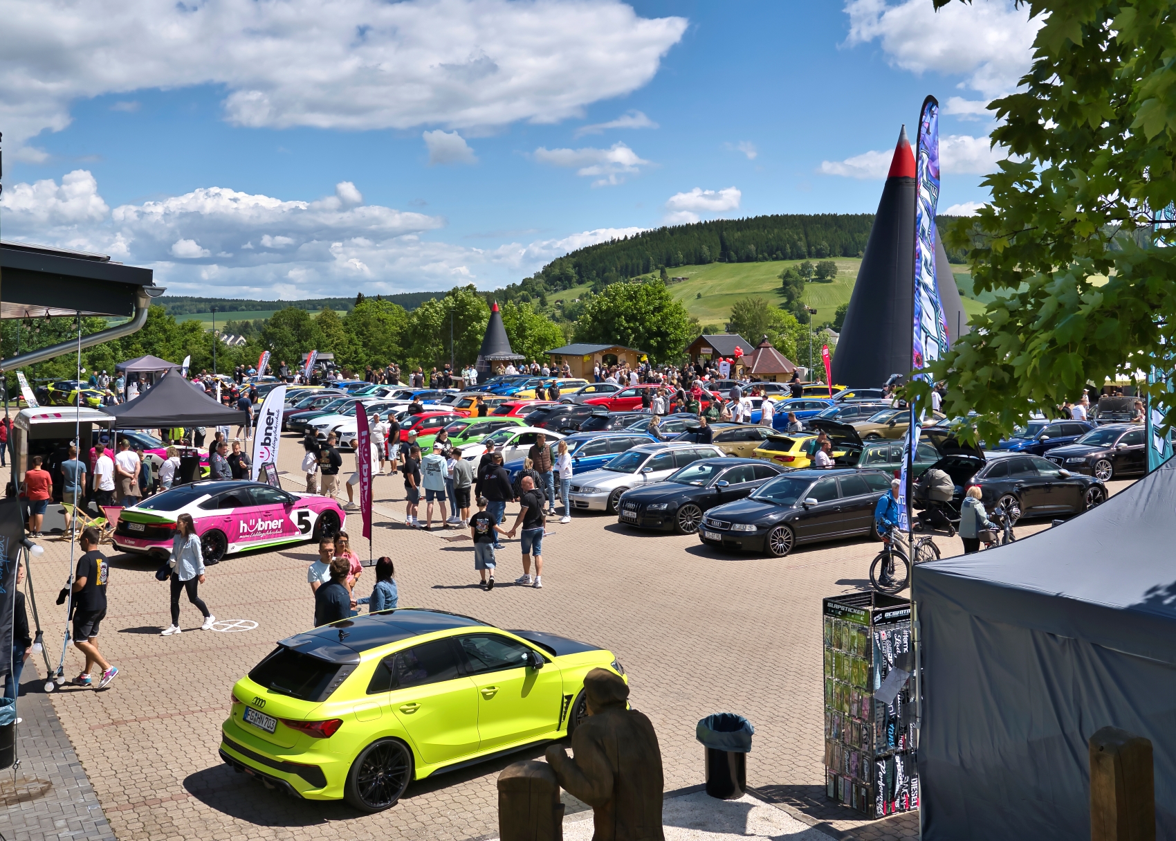 Besucher und viele Audi Fahrzeuge auf dem Eventgelände vor der großen Räucherkerze unter blauem Himmel.