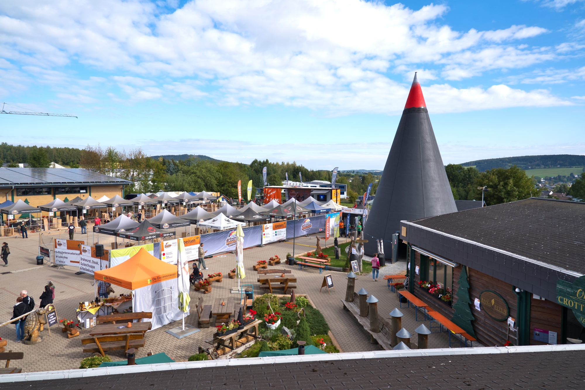 Übersicht über ein Veranstaltungsgelände im Freien mit zahlreichen Pavillons, Holzbänken und Besuchern, dominiert von einem großen schwarzen Kegelturm mit roter Spitze und Holzhütten im Vordergrund.